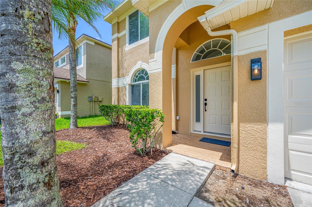 A beige house exterior features a covered entryway, a white front door, and landscaping — 25147 GEDDY DRIVE, LAND O LAKES, FL