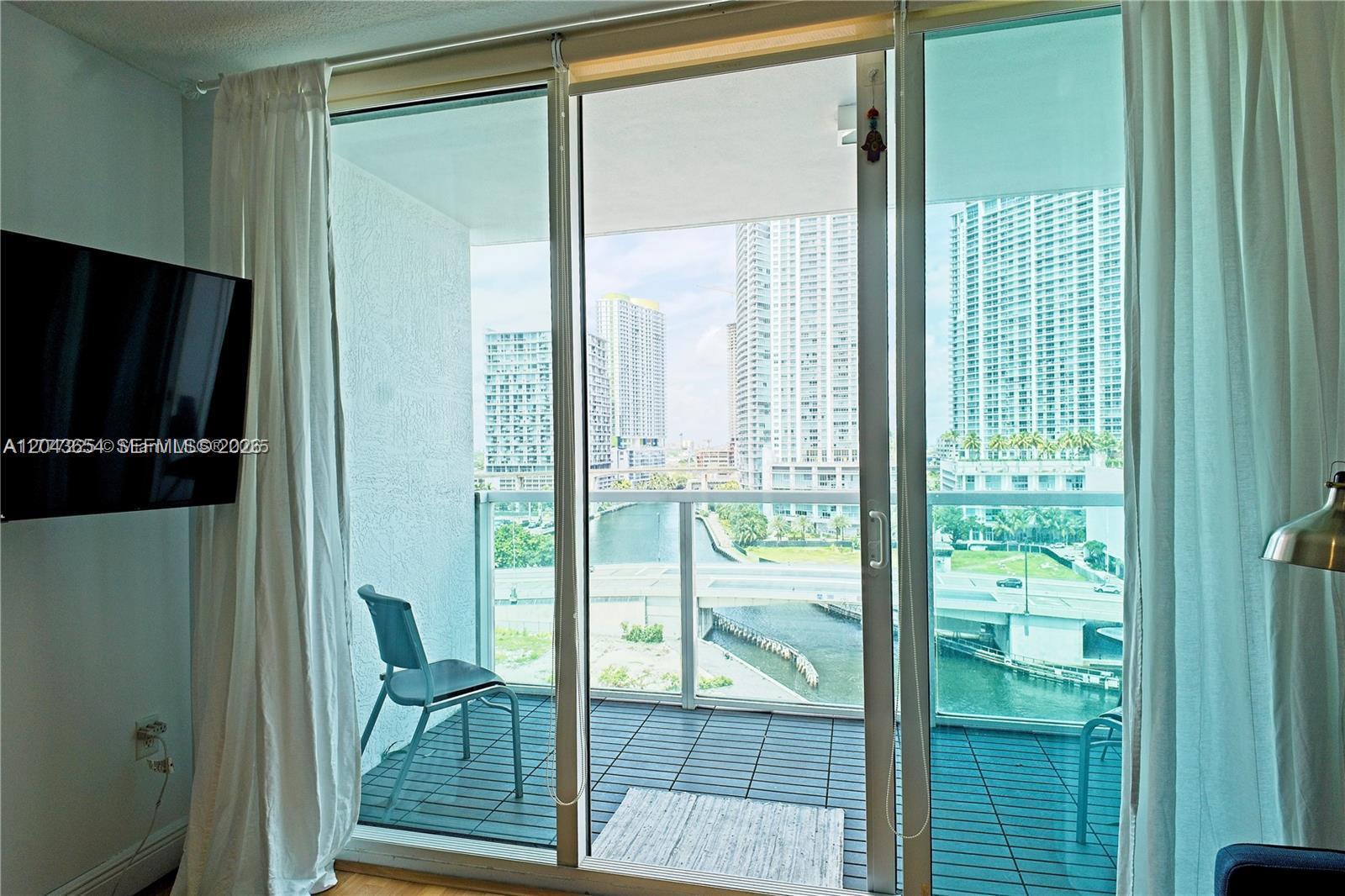 A room with a television and curtains opens onto a balcony overlooking a city canal — 31 SE 5th # 1203, Miami FL 33131, MIAMI, FL
