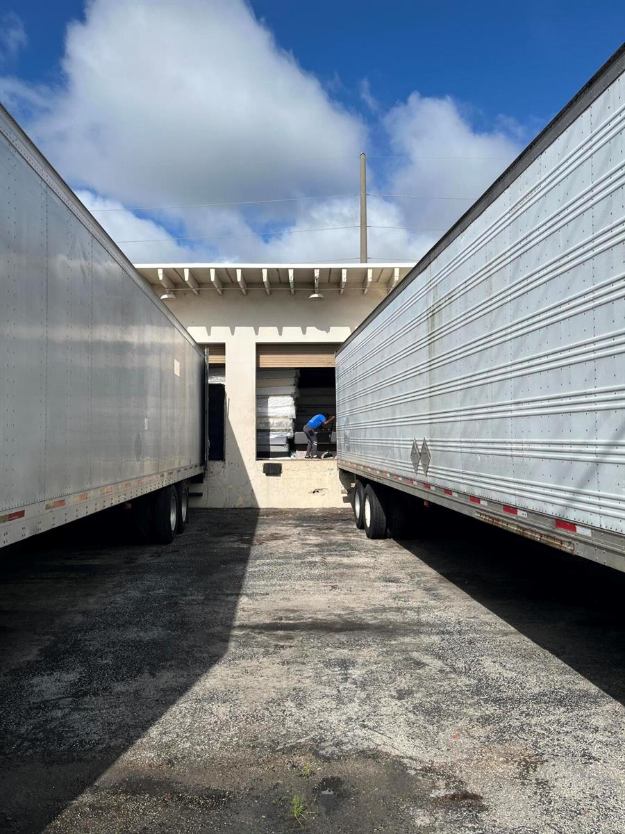 A person loads mattresses into a warehouse bay between two large parked semi-trailers — 5 NW 3rd Avenue, Dania Beach FL 33004, DANIA BEACH, FL