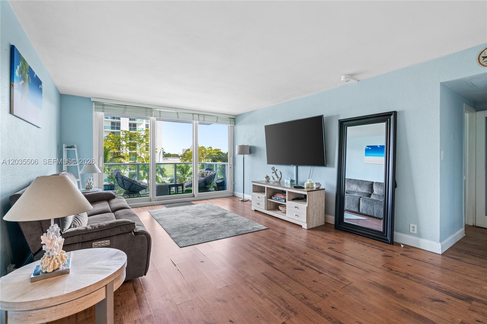 A living room features light blue walls, wood-look flooring, a large television, and balcony access — 1000 West Ave # 308, Miami Beach FL 33139, MIAMI BEACH, FL
