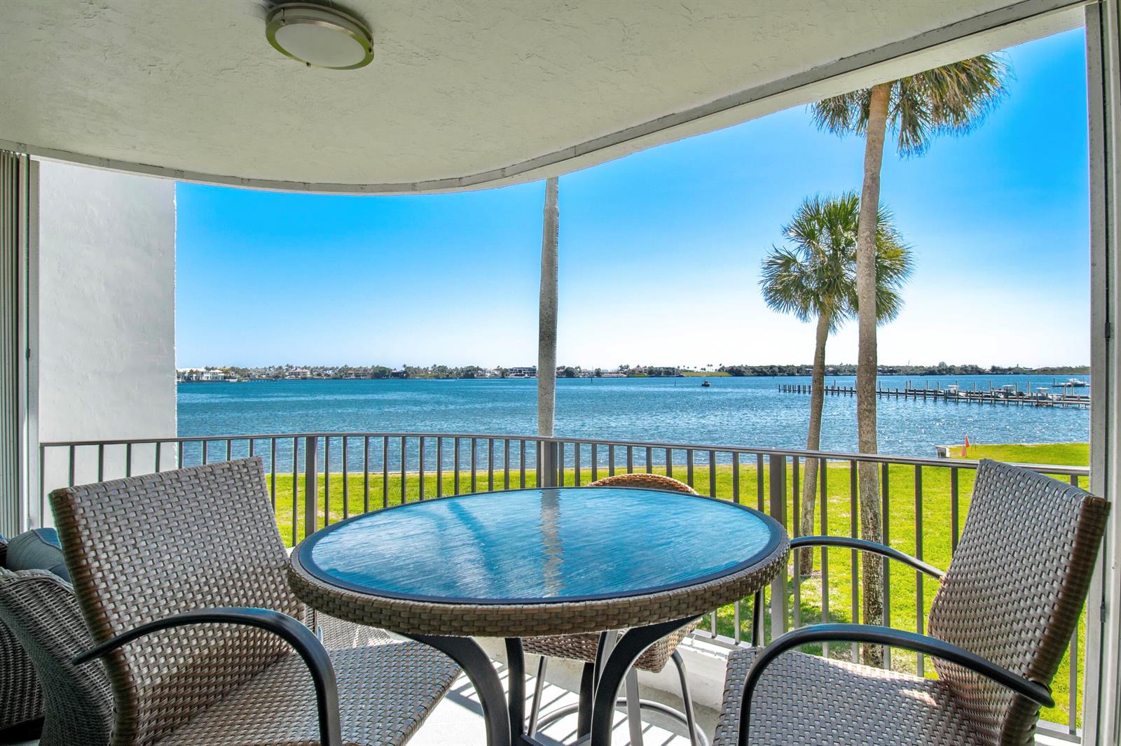 A balcony with wicker chairs and a glass table overlooks a bay with palm trees — 8200 Lakeshore Drive # 202, Hypoluxo FL 33462, HYPOLUXO, FL