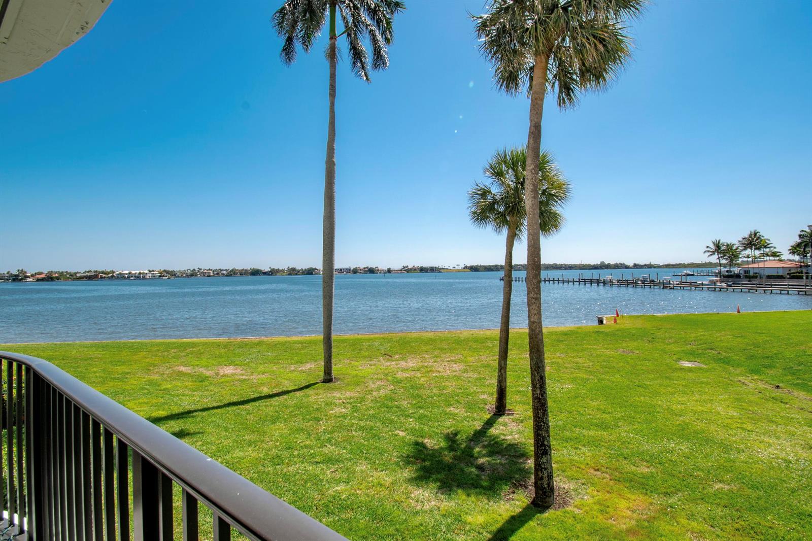 A view from a balcony overlooking a grassy lawn, palm trees, and a calm bay — 8200 Lakeshore Drive # 202, Hypoluxo FL 33462, HYPOLUXO, FL