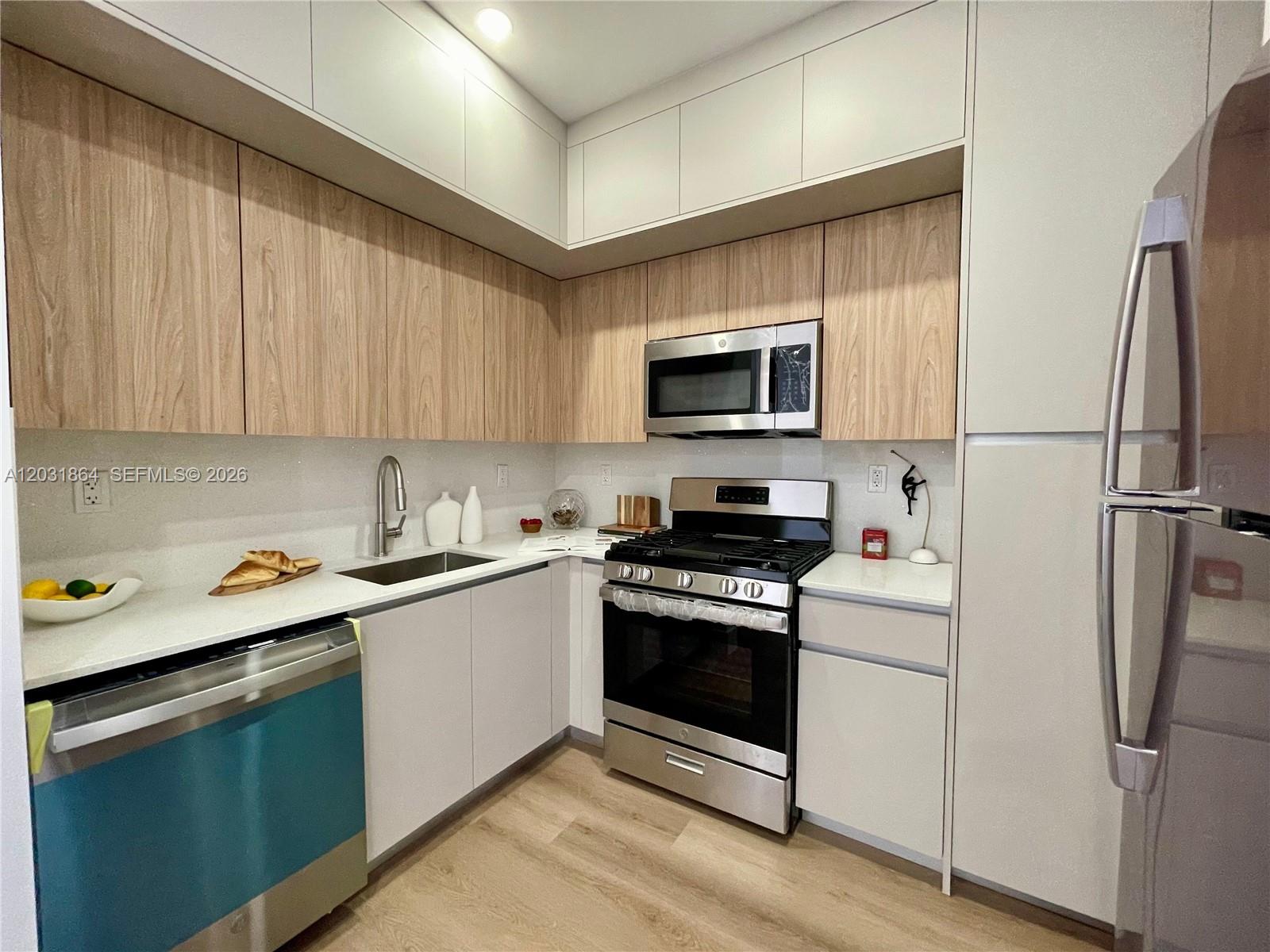 A modern kitchen features light wood-grain upper cabinets, white lower cabinets, and stainless steel appliances — 1619 Jefferson Ave # 107, Miami Beach FL 33139, MIAMI BEACH, FL