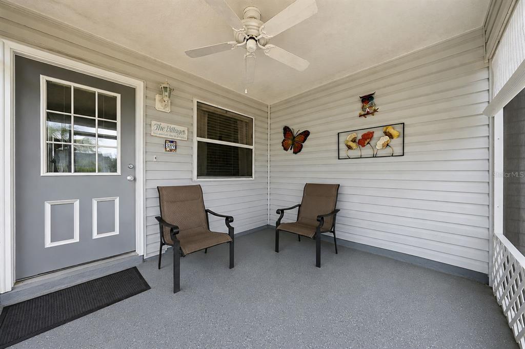 A covered porch features a gray door, two chairs, a ceiling fan, and wall decorations — 389 SEVEN OAKS LANE, THE VILLAGES, FL