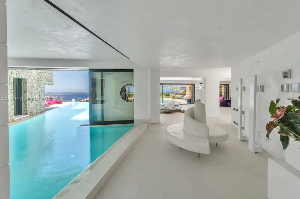 A modern living area features a curved white sofa and an indoor-outdoor infinity pool — Sardinia Luxury Estate Porto Cervo Max Mlekus, FL