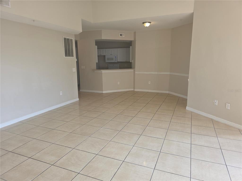 An empty room with beige tile flooring and a pass-through opening to a kitchen — 5566 METROWEST BOULEVARD UNIT 303, ORLANDO, FL