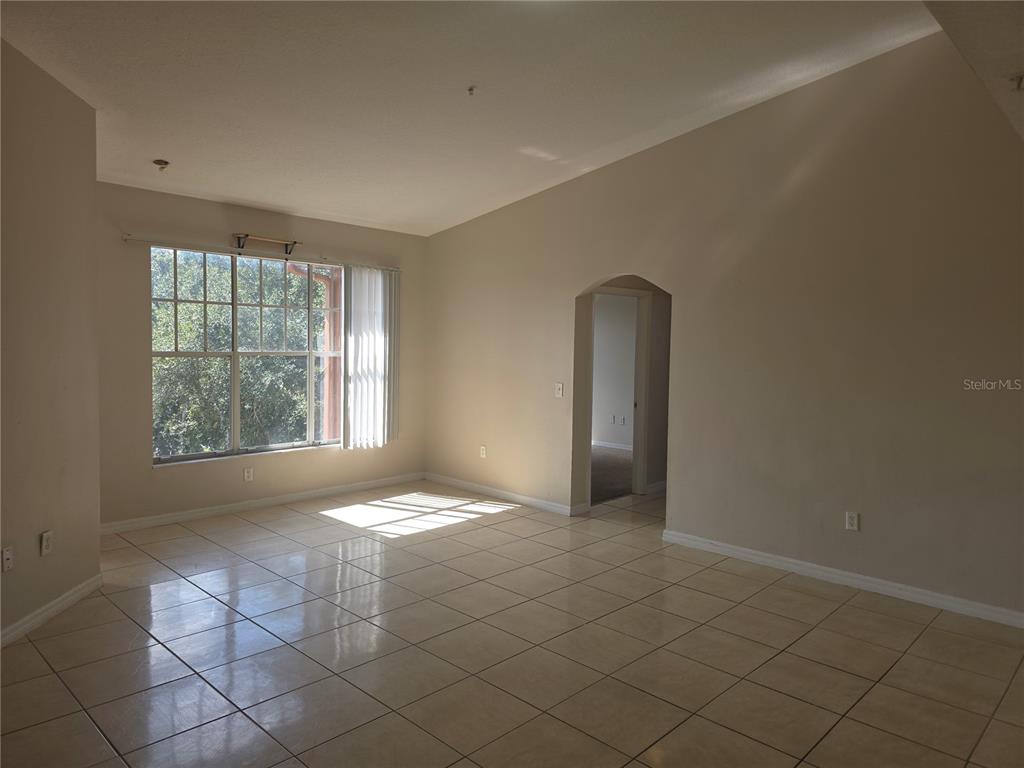 An empty living room features beige tile flooring, vaulted ceilings, and a large window — 5566 METROWEST BOULEVARD UNIT 303, ORLANDO, FL