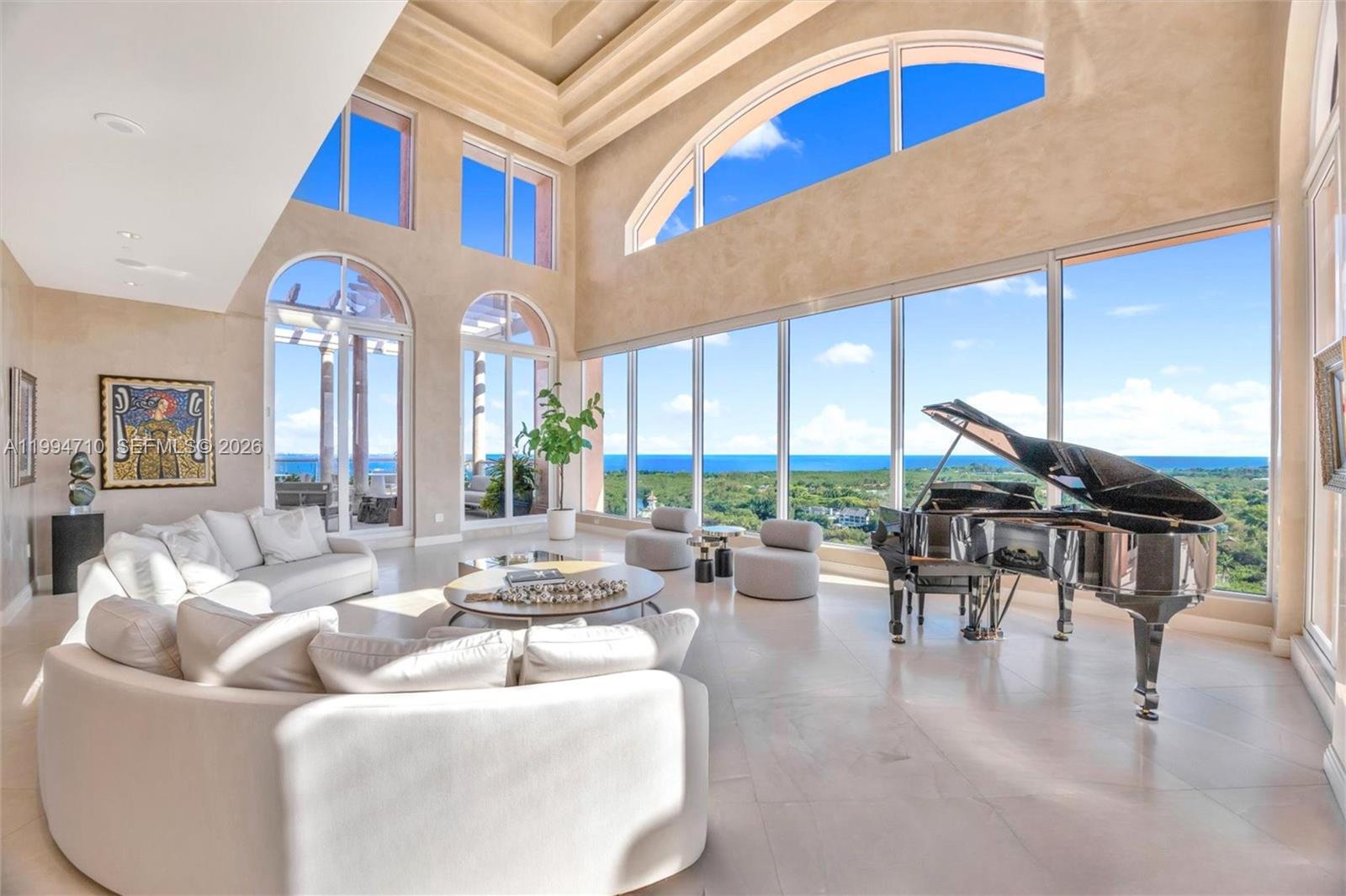 A spacious living room features a grand piano, white sofas, and floor-to-ceiling windows with ocean views — 60 Edgewater Dr # A, Coral Gables FL 33133, CORAL GABLES, FL