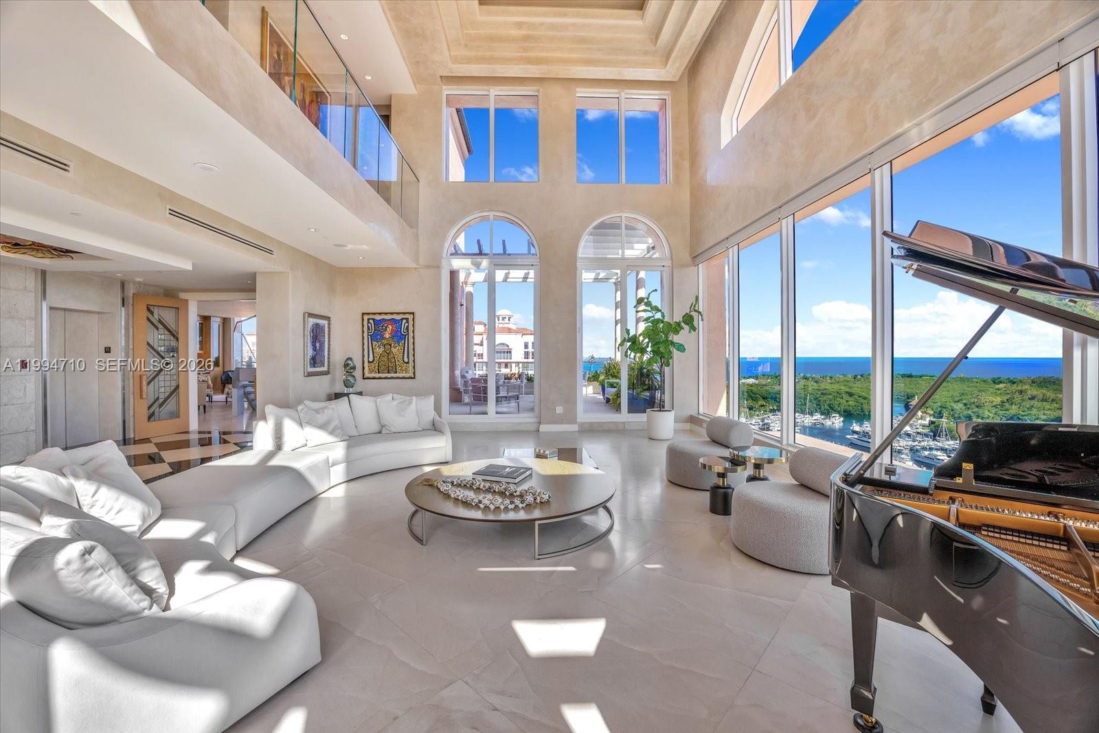 A spacious living room with high ceilings, a grand piano, and floor-to-ceiling windows overlooking water — 60 Edgewater Dr # A, Coral Gables FL 33133, CORAL GABLES, FL