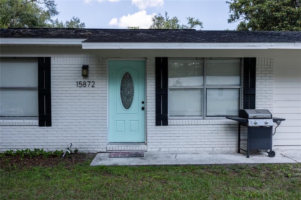 A white brick house exterior features a light blue front door and a charcoal grill — 15872 SW 133RD LANE, ARCHER, FL