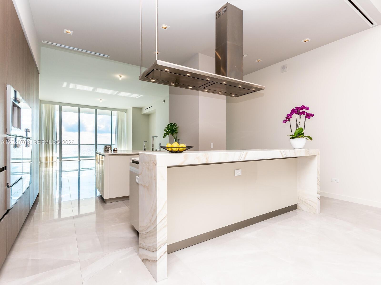 A modern kitchen features a marble waterfall island, stainless steel range hood, and tile flooring — 18501 E Collins # 1503, Sunny Isles Beach FL 33160, SUNNY ISLES BEACH, FL