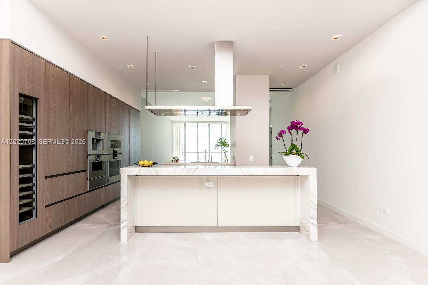 A modern kitchen features a marble-topped island, wood cabinetry, and a stainless steel range hood — 18501 E Collins # 1503, Sunny Isles Beach FL 33160, SUNNY ISLES BEACH, FL