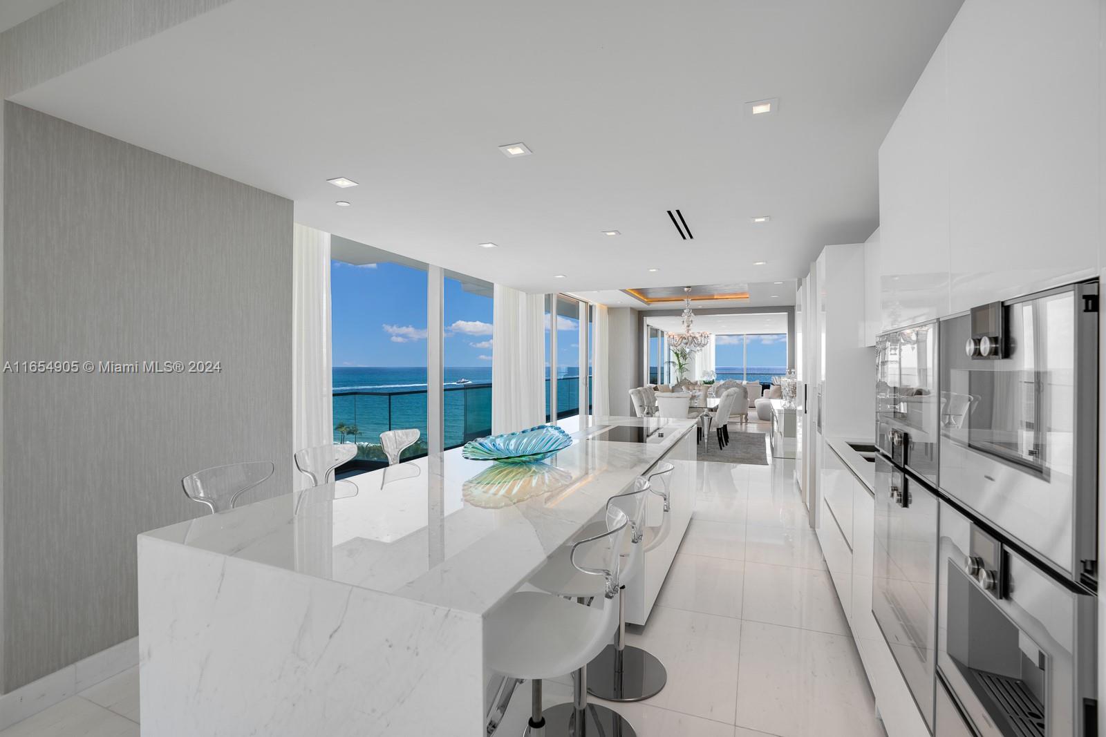 A modern kitchen features a marble island, white cabinetry, and floor-to-ceiling windows overlooking the ocean — 10203 Collins Ave # 801, Bal Harbour FL 33154, BAL HARBOUR, FL