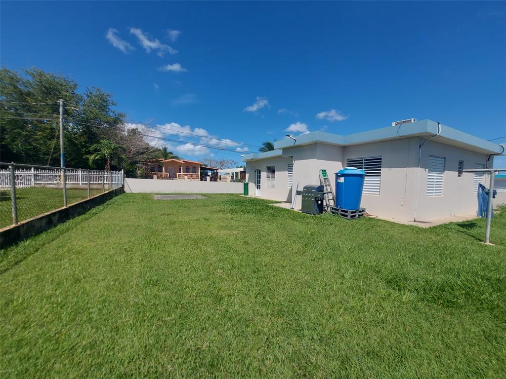 A grassy backyard features a concrete house, a blue water tank, and a metal fence — Carr. 459 BARRIO CEIBA ALTA, AGUADILLA, PR