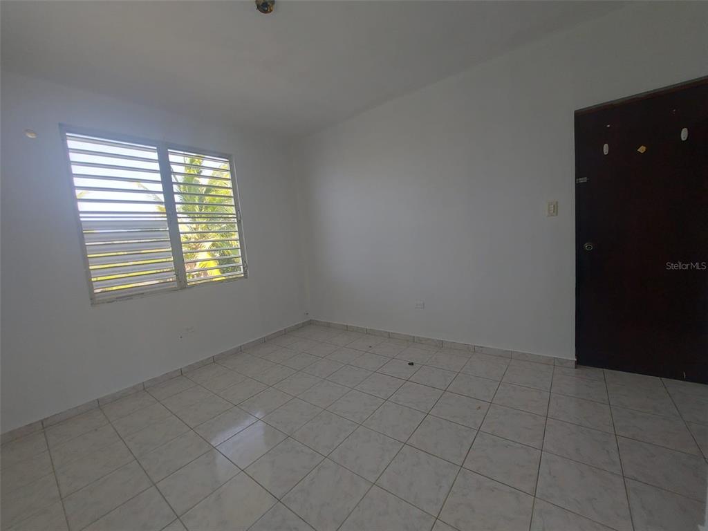 An empty room features white walls, light-colored tiled flooring, a louvered window, and a dark door — Carr. 459 BARRIO CEIBA ALTA, AGUADILLA, PR