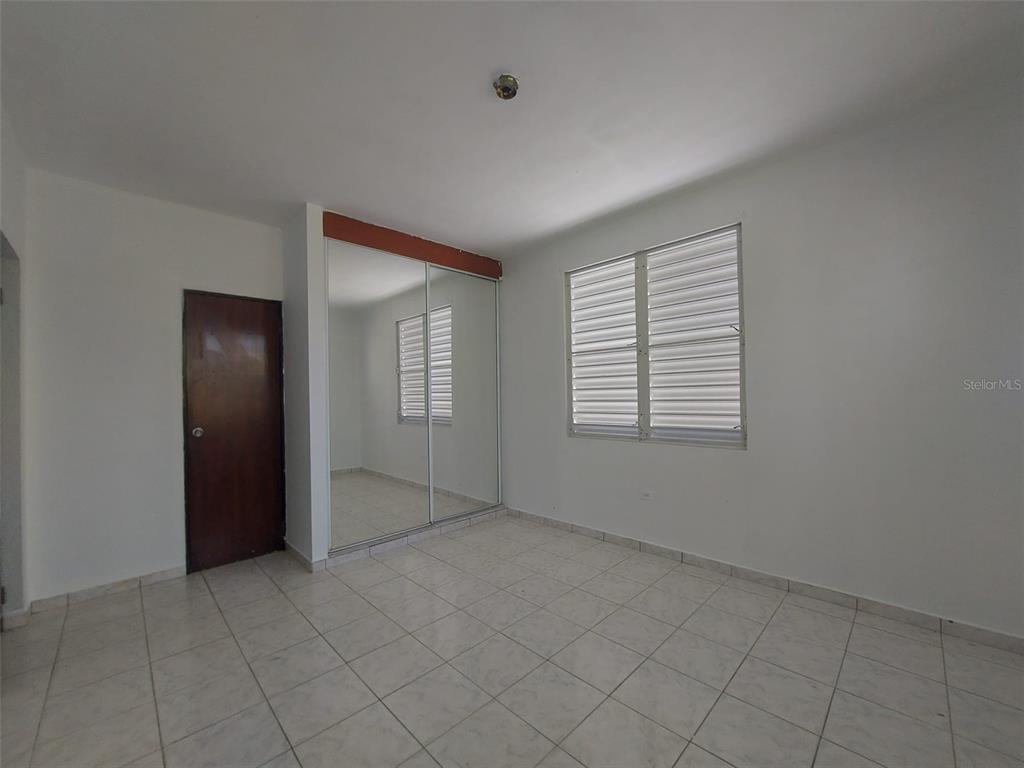 A bedroom with white walls, tiled floors, a dark wood door, and mirrored closet doors — Carr. 459 BARRIO CEIBA ALTA, AGUADILLA, PR
