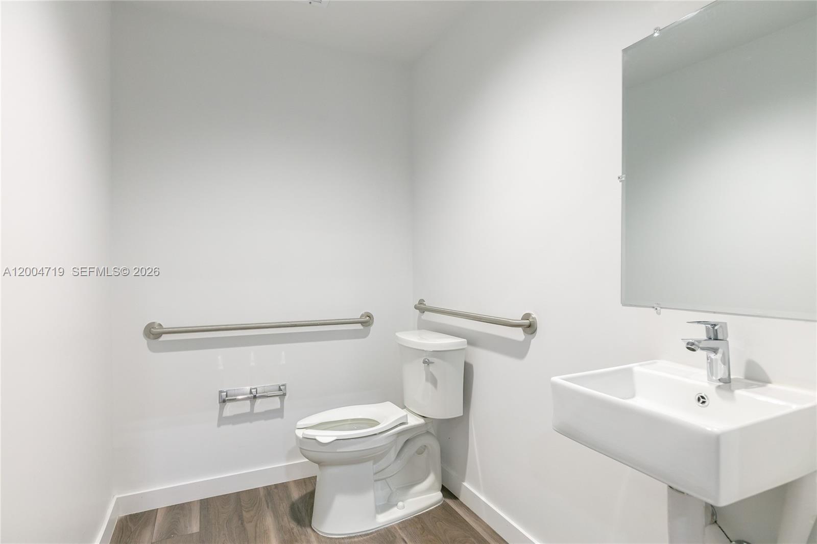 A minimalist bathroom features a white toilet, a wall-mounted sink, and ADA-compliant grab bars — 7265 NW 74th St # 6, Medley FL 33166, MEDLEY, FL