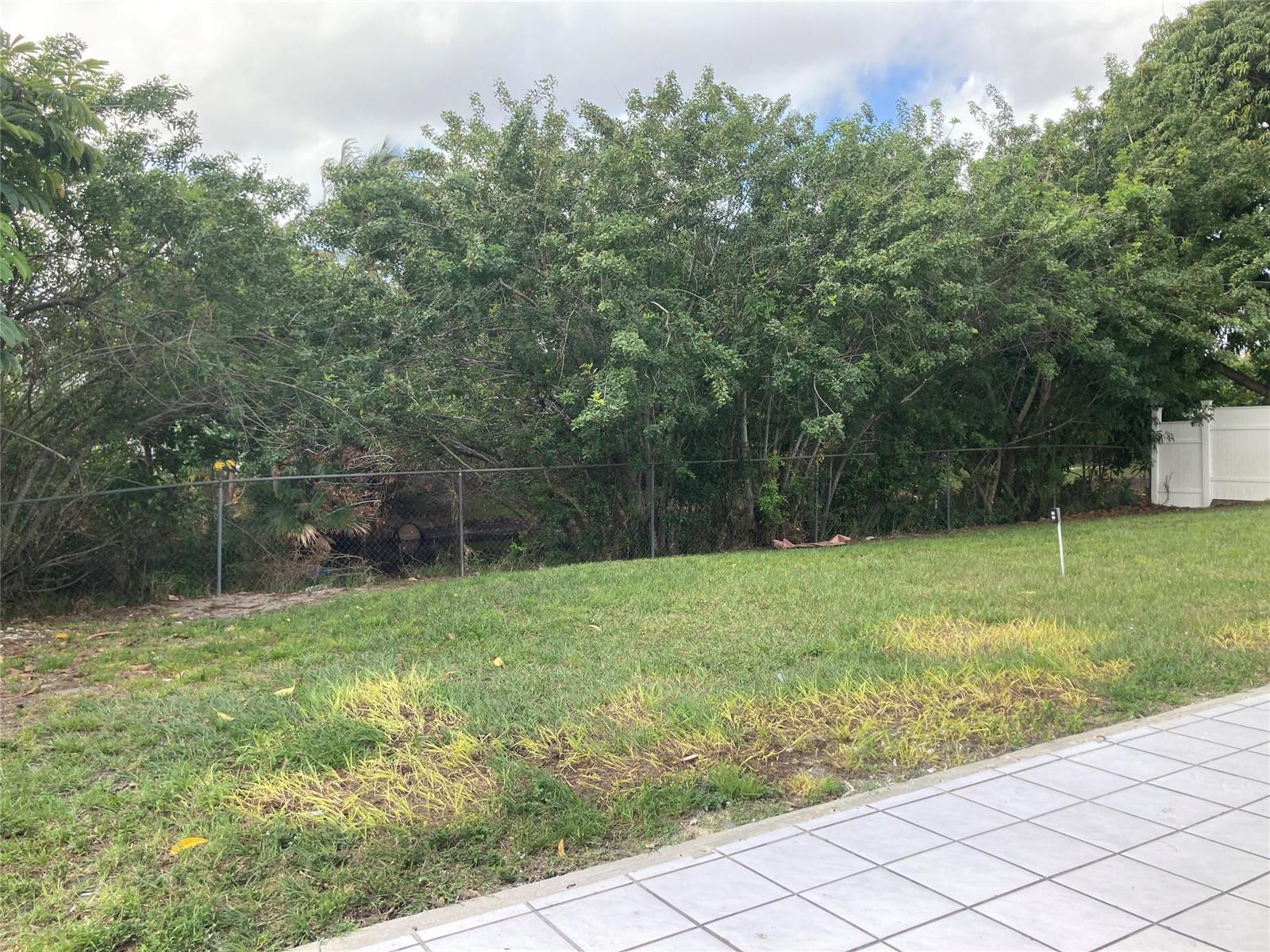A grassy backyard features a chain-link fence bordering dense trees and a white privacy fence — 6230 SW 4th Place, Margate FL 33068, MARGATE, FL