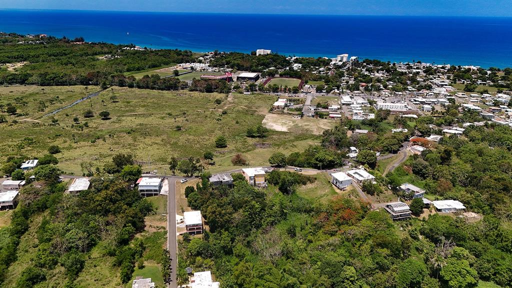 An aerial view shows a coastal town with scattered houses, green fields, and the ocean — CARR 115 KM HM 11.3, RINCON, PR