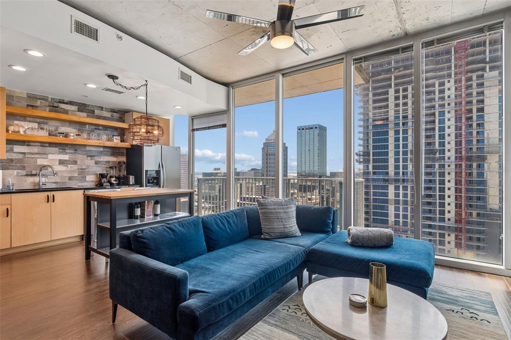 A modern living room with a blue sectional sofa, kitchen area, and floor-to-ceiling city views — 777 N ASHLEY DRIVE UNIT 2308, TAMPA, FL