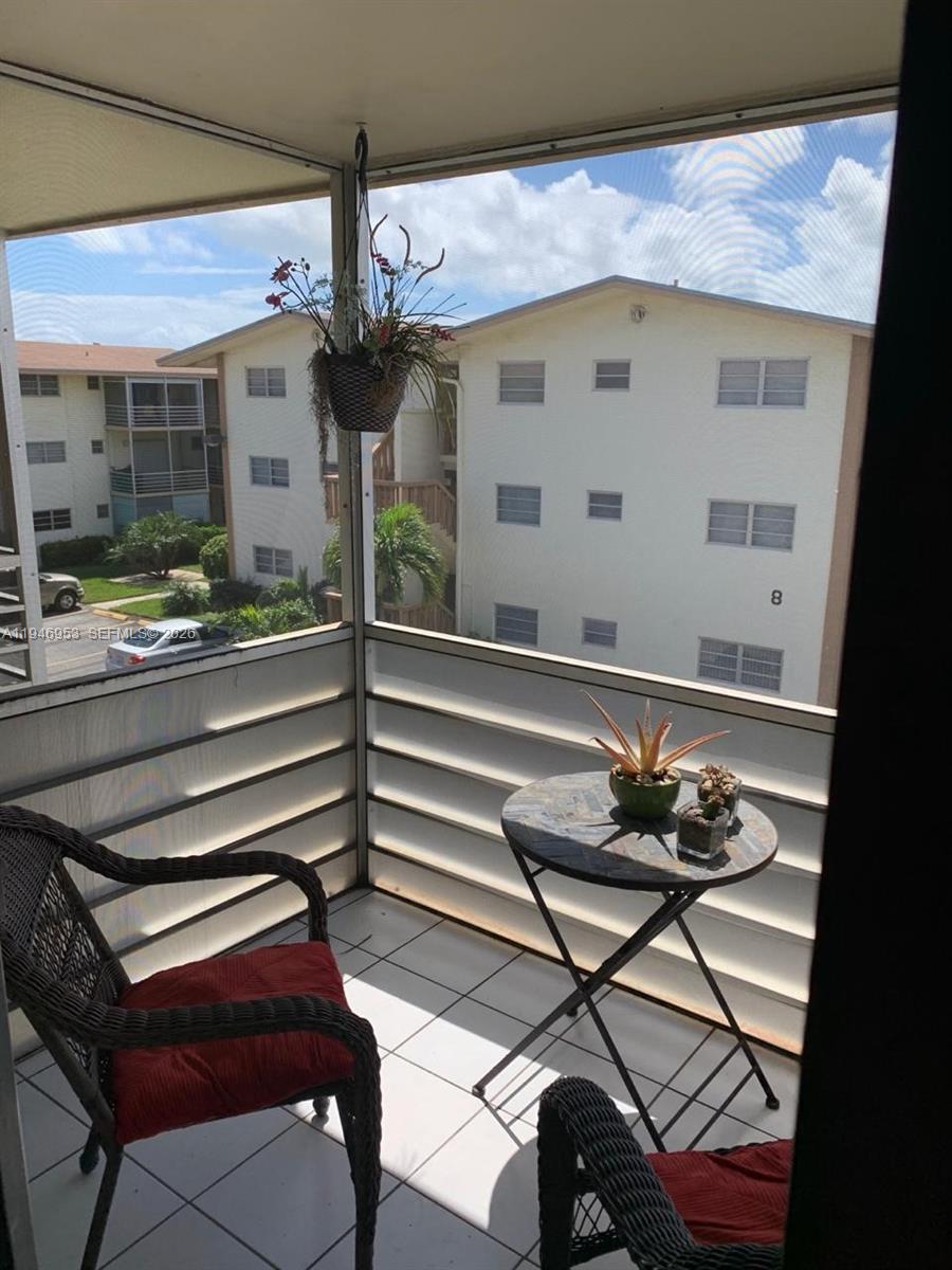 A screened-in balcony features white tiled flooring, two wicker chairs, and a small round table — 16801 NE 13th Ave # 301, Miami FL 33162, MIAMI, FL