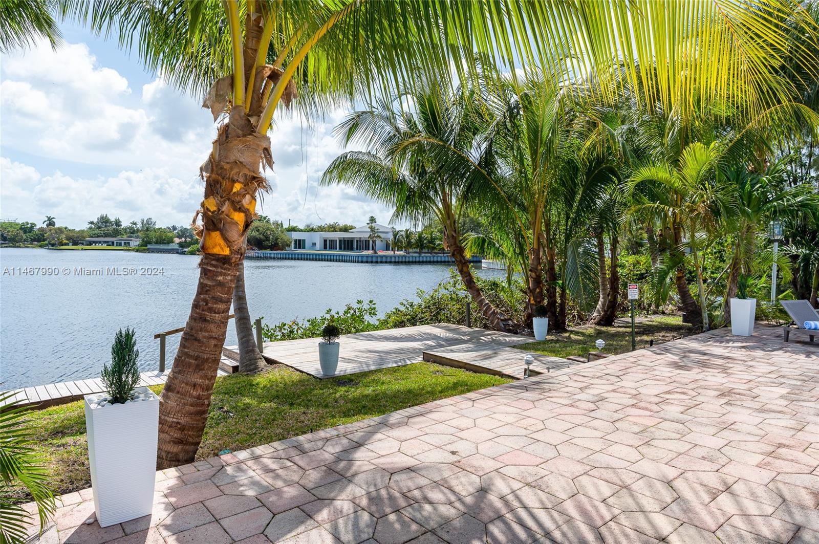 A paved patio overlooks a canal with a wooden dock and several palm trees — 2500 NE 199th St, Miami FL 33180, MIAMI, FL