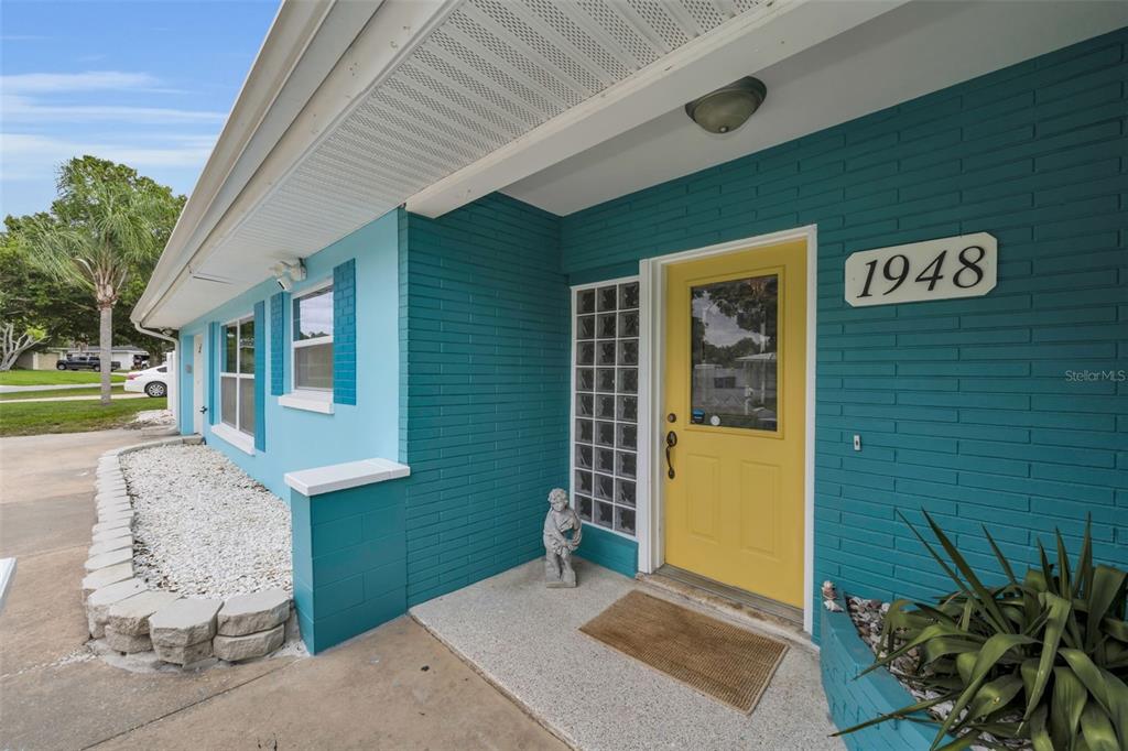 A teal brick house exterior features a yellow front door and a glass block window — 1948 BYRAM DRIVE, CLEARWATER, FL
