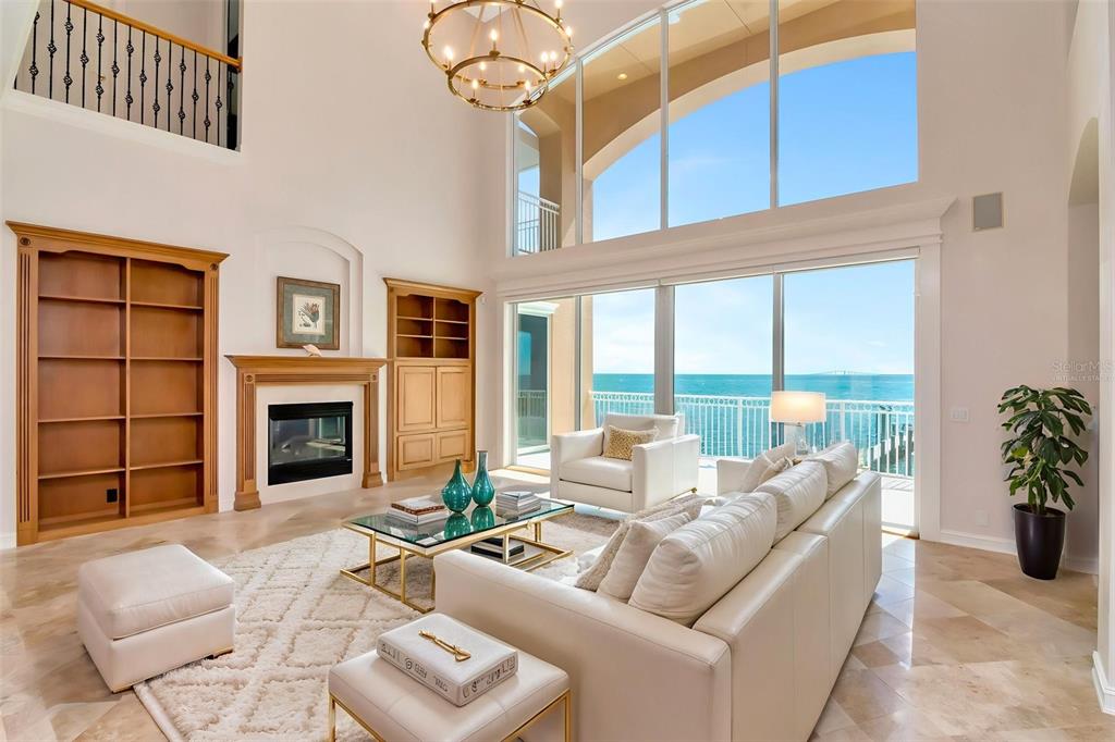 A high-ceilinged living room features a fireplace, built-in shelving, and floor-to-ceiling windows overlooking the ocean — 7680 PARADISE POINTE CIRCLE S, ST PETERSBURG, FL