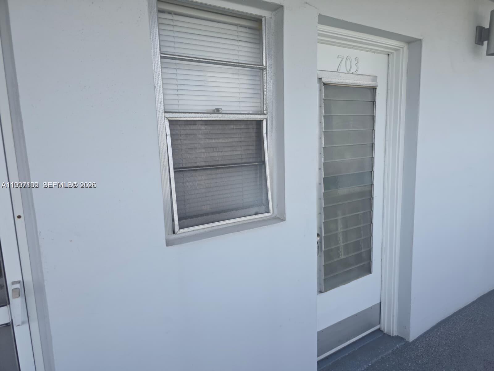 A white exterior wall features a window with blinds and a door labeled 703 — 320 NE 12th Ave # 703, Hallandale Beach FL 33009, HALLANDALE BEACH, FL