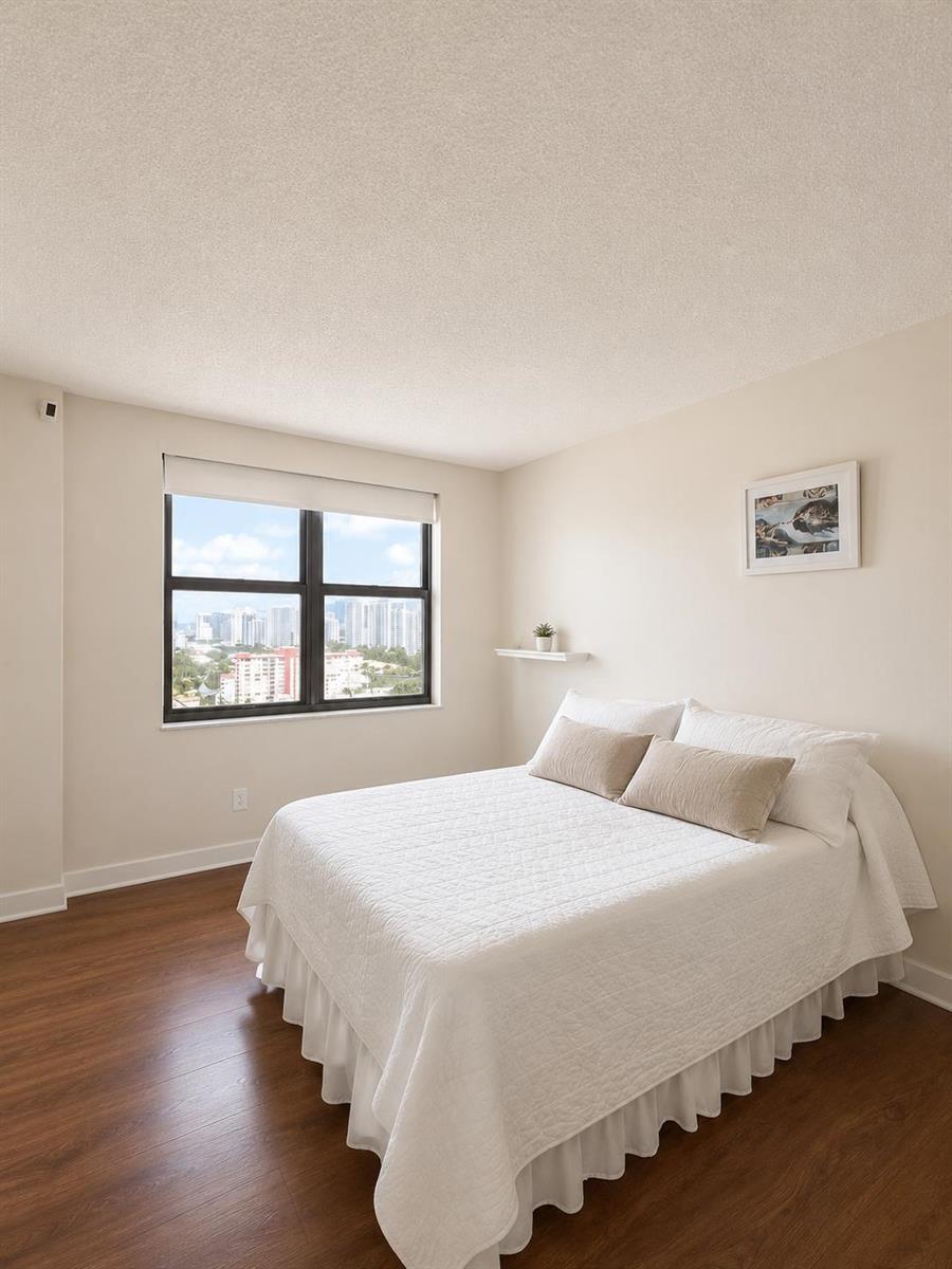 A bedroom with a white bed, wood-look flooring, and a window overlooking a city skyline — 1985 S Ocean Drive # 22n, FL 33009, FL
