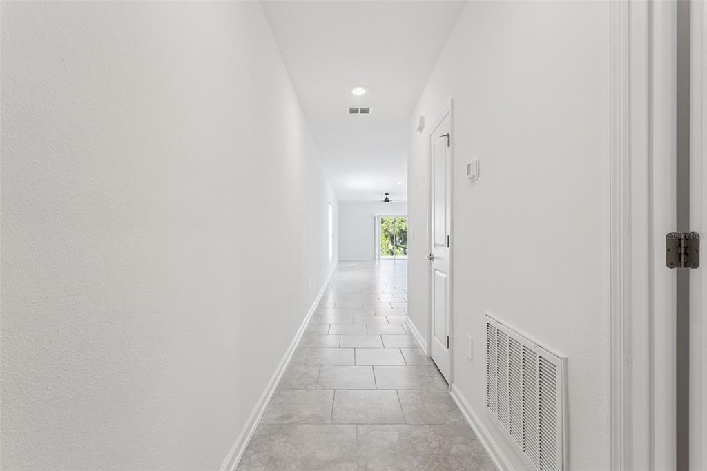 A long, white-walled hallway with light gray tile flooring leads toward a bright living area — 26180 FEATHERGRASS CIRCLE, LEESBURG, FL