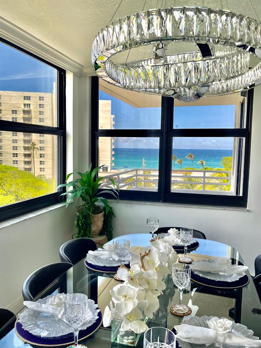 A dining room with a glass table, crystal chandelier, and ocean views through large windows — 3450 S Ocean Boulevard # 605, Highland Beach FL 33487, HIGHLAND BEACH, FL