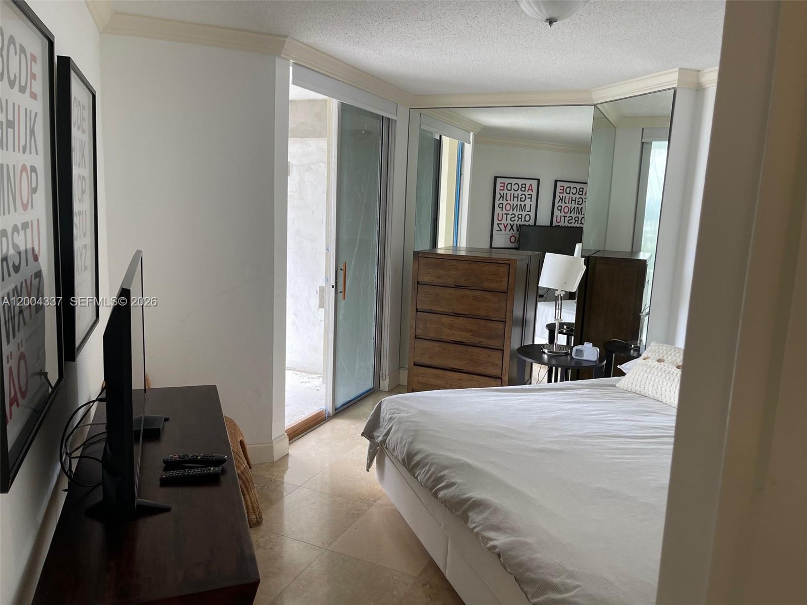 A bedroom features a bed, a wooden dresser, a television, and a sliding glass door — 5555 Collins Ave # 7Z, Miami Beach FL 33140, MIAMI BEACH, FL
