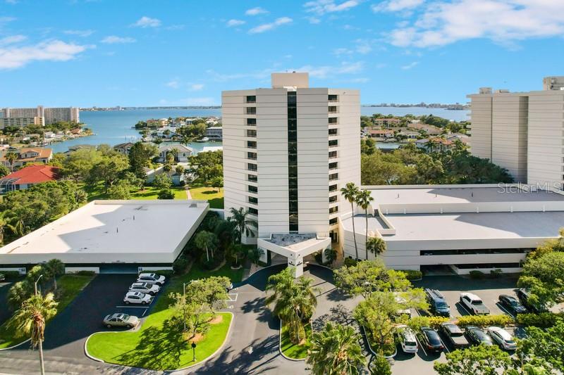 An aerial view shows a tall, white residential building surrounded by parking lots and lush greenery — 5940 PELICAN BAY PLAZA S UNIT 1105, GULFPORT, FL