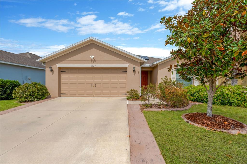 A tan single-story house features a two-car garage, a front walkway, and a large tree — 8529 HUNTERS FORK LOOP, RUSKIN, FL