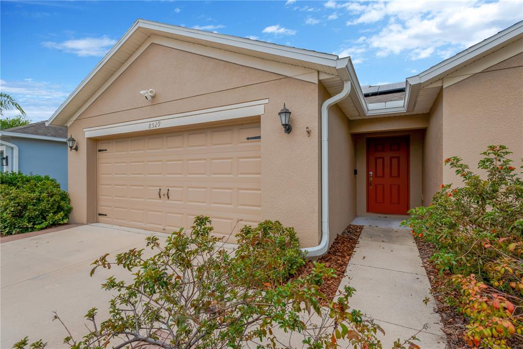 A tan house exterior features a two-car garage, a red front door, and landscaping — 8529 HUNTERS FORK LOOP, RUSKIN, FL