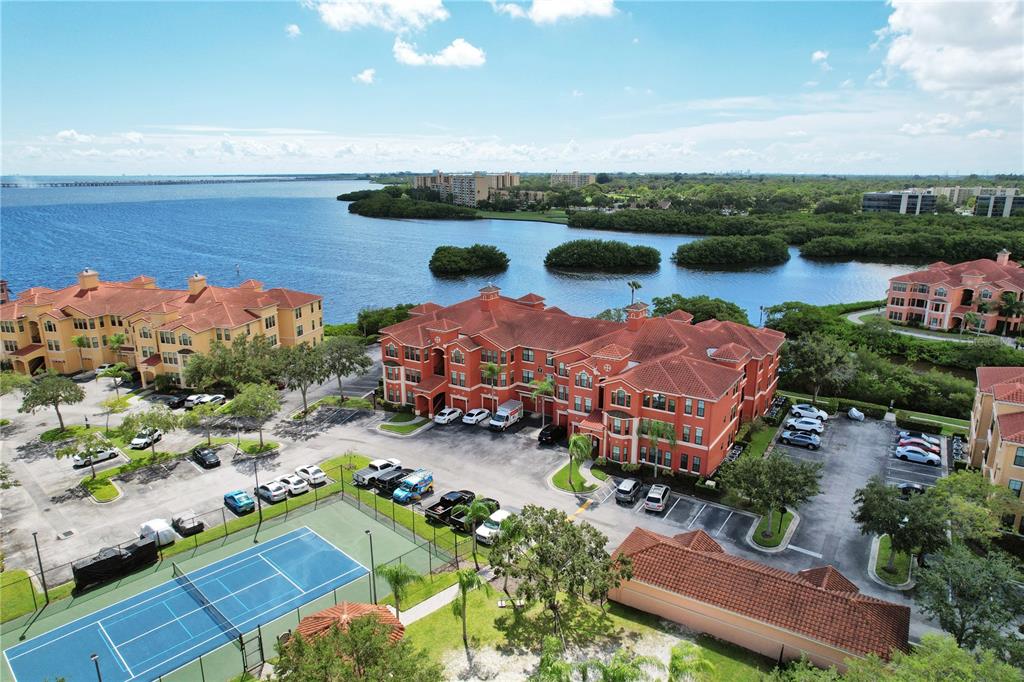 An aerial view shows apartment buildings with red tile roofs, a tennis court, and water — 2724 VIA MURANO UNIT 638, CLEARWATER, FL