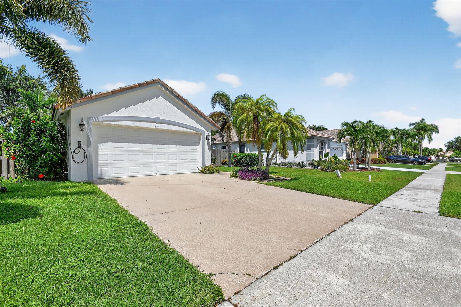 A single-story house with a white garage, concrete driveway, and palm trees under a blue sky — 1891 Oak Berry Circle, Wellington FL 33414, WELLINGTON, FL