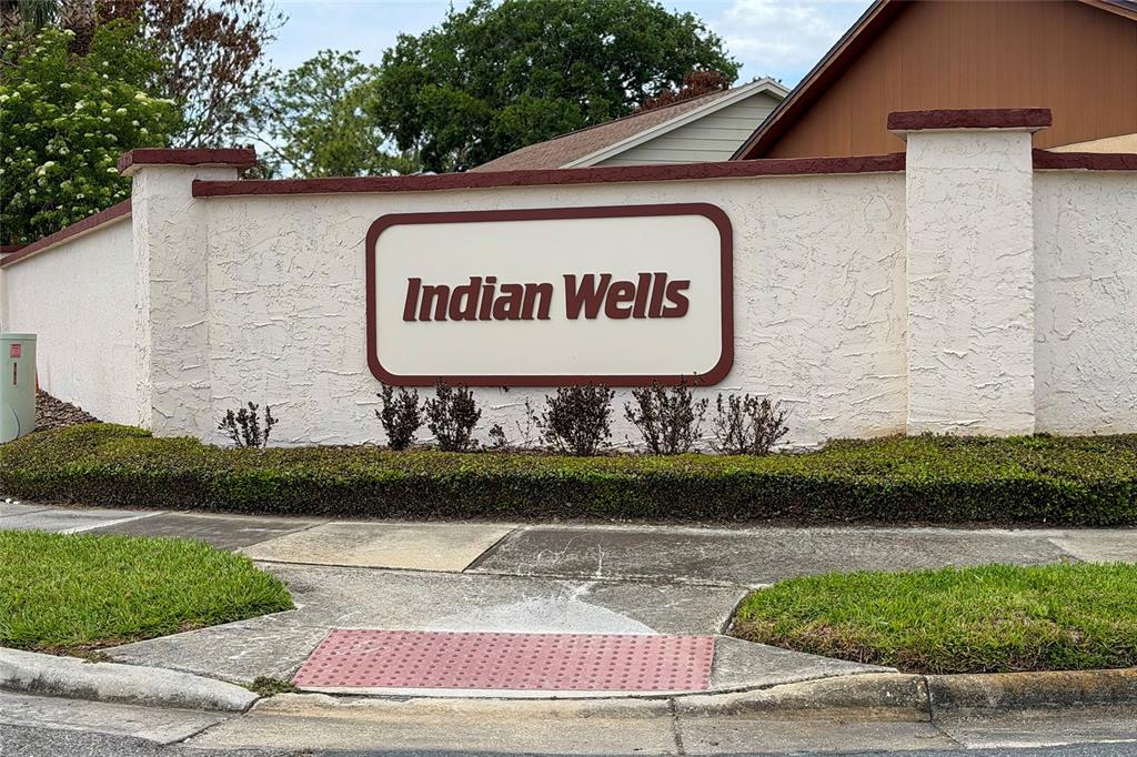 A white stucco wall features a sign reading Indian Wells above a manicured hedge — 4980 WARRIOR LANE, KISSIMMEE, FL