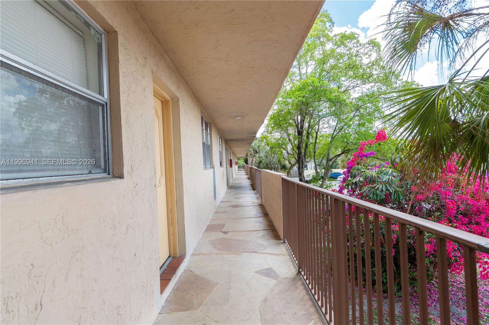 An outdoor apartment walkway features a tan stucco wall, a metal railing, and stone flooring — 3900 NW 76th Ave # 212, Sunrise FL 33351, SUNRISE, FL