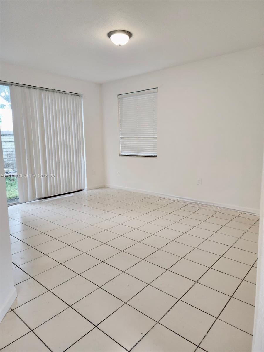 An empty room features white walls, light-colored floor tiles, a sliding glass door, and blinds — HOMESTEAD, FL