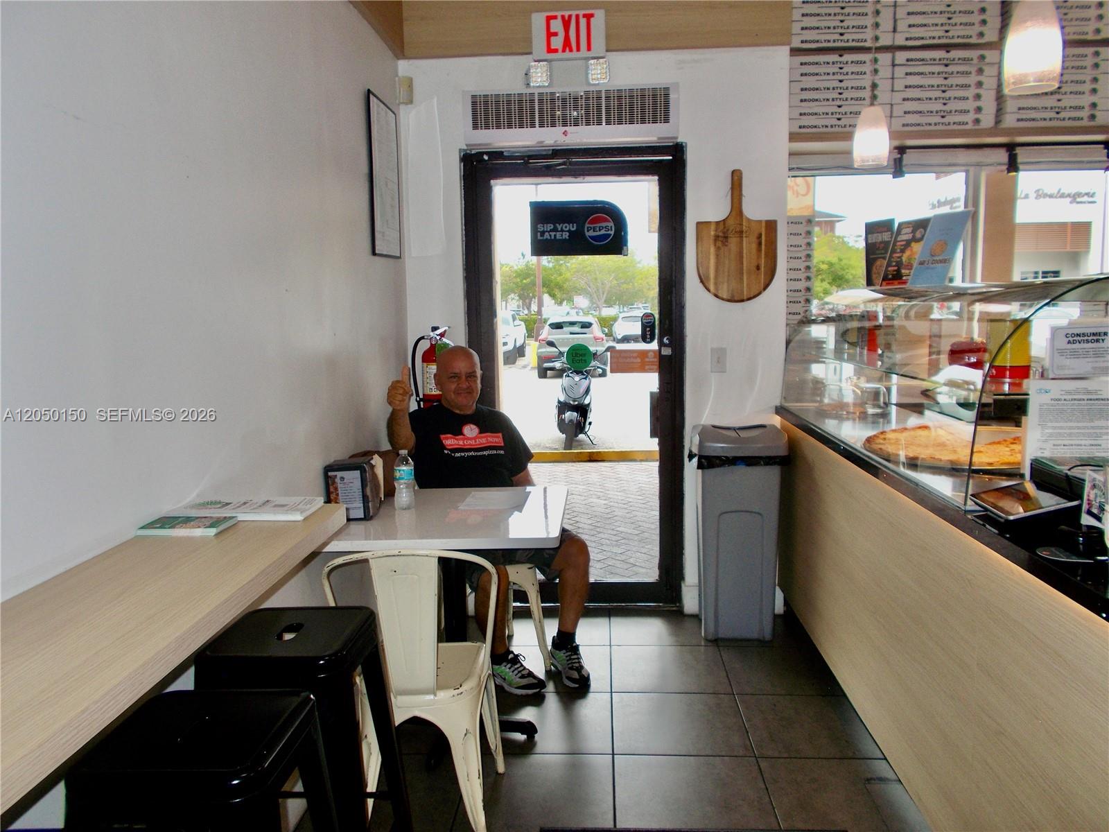 A man sits at a table inside a pizza shop with a glass display case — 8237 SW 124th St, Pinecrest FL 33156, PINECREST, FL