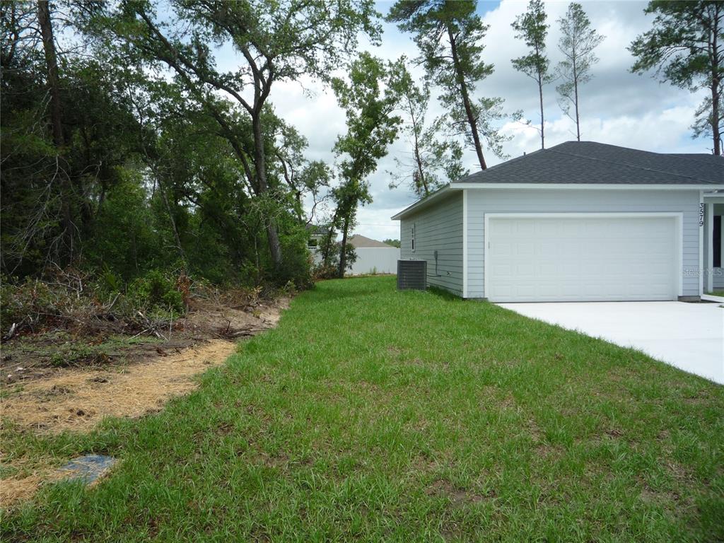 A side view of a house exterior showing a garage, lawn, and adjacent wooded area — 3579 SW 128TH PL, OCALA, FL