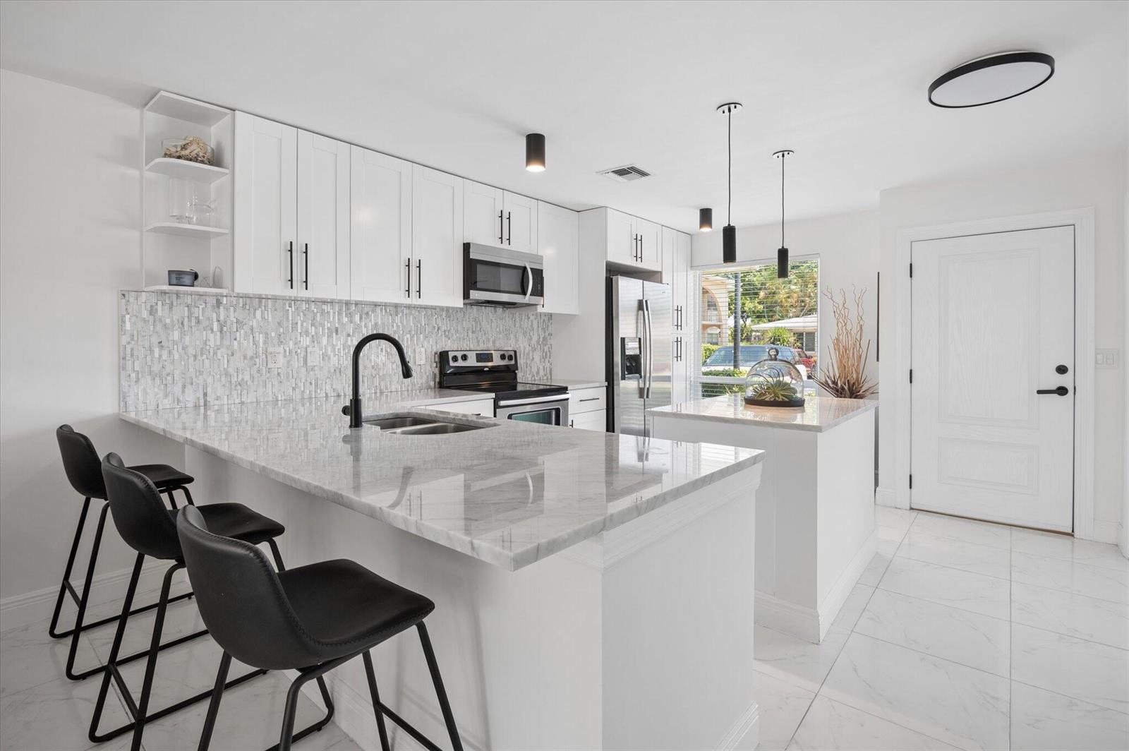 A modern white kitchen features marble countertops, a breakfast bar with stools, and stainless appliances — 13725 Flora Place # C, Delray Beach FL 33484, DELRAY BEACH, FL