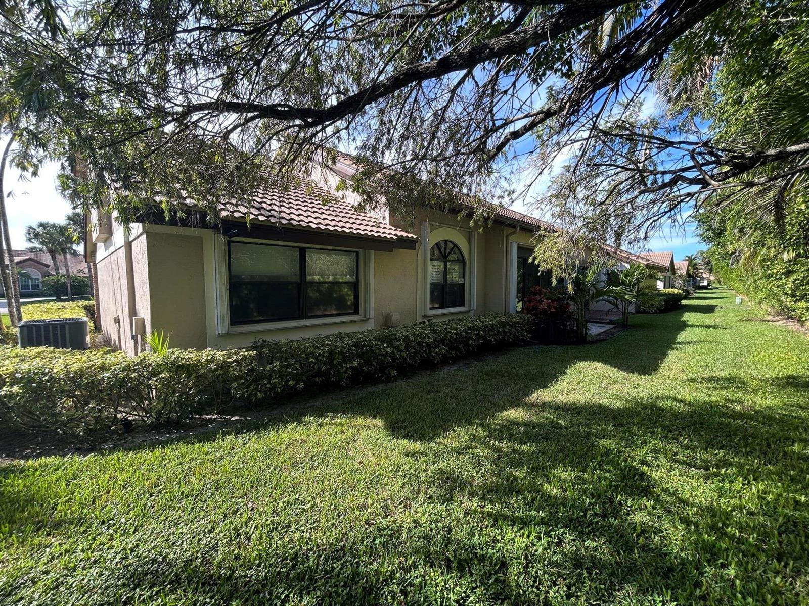 A side view of a tan stucco house with a tiled roof and landscaped yard — 7207 Le Chalet Boulevard, Boynton Beach FL 33472, BOYNTON BEACH, FL