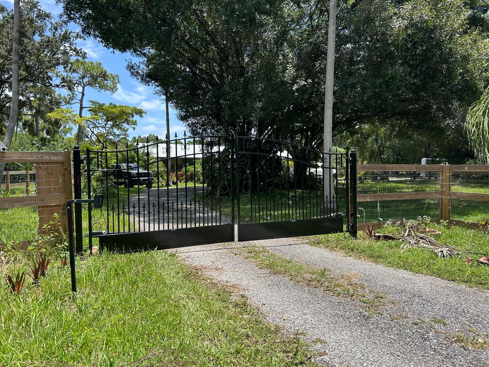 A black metal driveway gate stands between wooden fence sections on a gravel path — 3208 A Road, FL 33470, FL