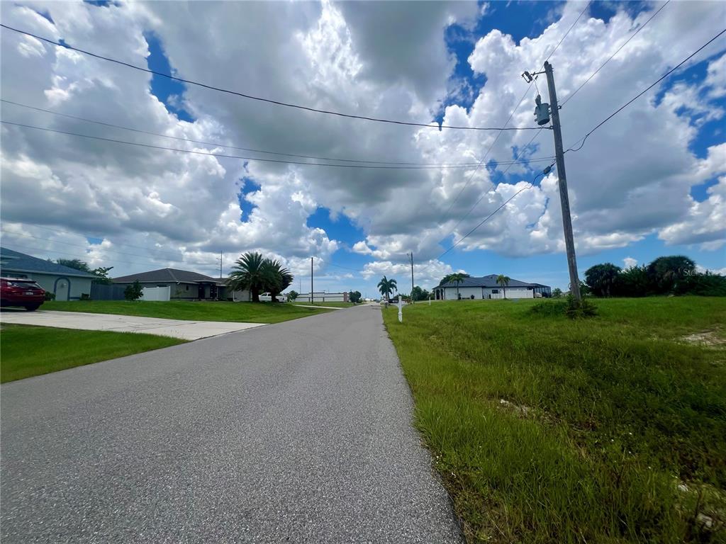 A paved residential street stretches forward under a cloudy blue sky with houses on both sides — 1423 NW 8TH TERRACE, CAPE CORAL, FL