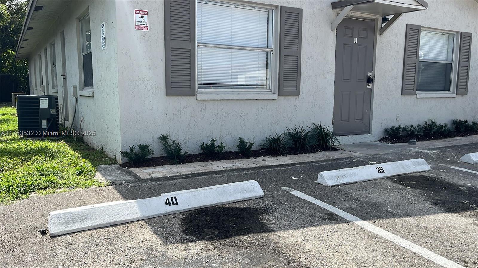 The exterior of a single-story building features a gray door, windows with shutters, and parking — 550 NE 44th St # M2, Deerfield Beach FL 33064, DEERFIELD BEACH, FL