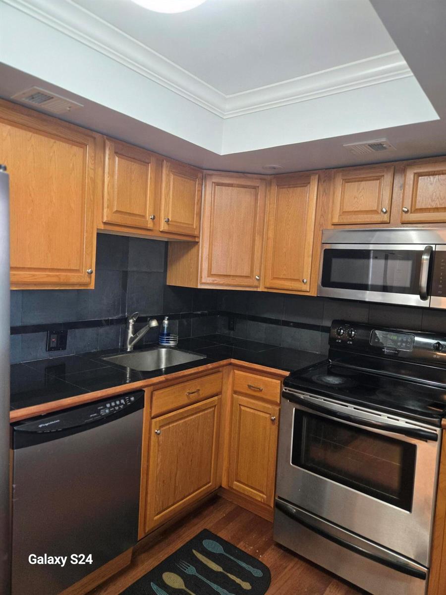 A kitchen features wood cabinets, black countertops, a dark tile backsplash, and stainless steel appliances — 2651 S Course Drive # 509, Pompano Beach FL 33069, POMPANO BEACH, FL