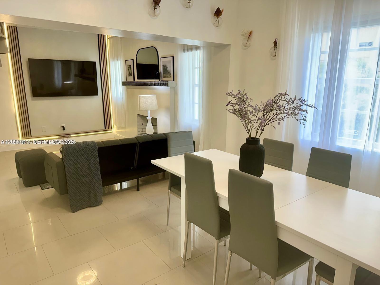 A dining area with a white table and grey chairs sits before a living room — 6966 Byron Ave # 101, Miami Beach FL 33141, MIAMI BEACH, FL